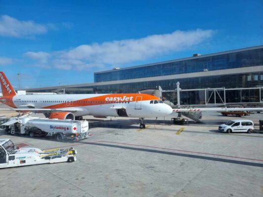 An Easyjet plane stopped near the terminal, at Malaga Airport. The plane is white with a large orange stripe on it, and an orange tail.