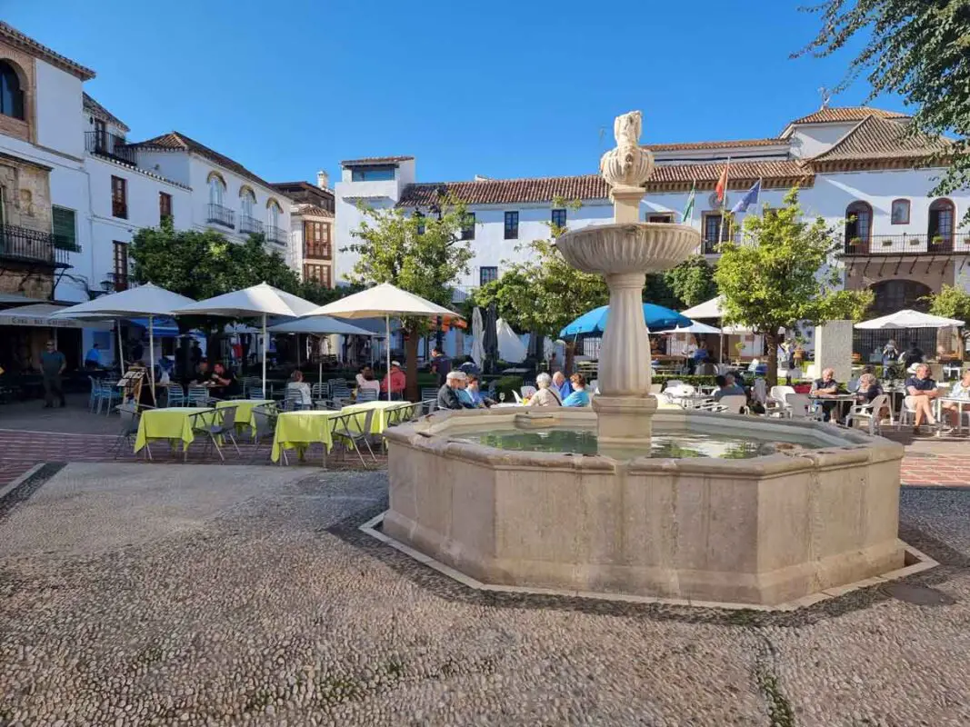 A round stone fountain in Plaza de los Naranjos in the centre of the Marbellaold town. The square is small and it is lined with orange trees in front of the white buildings. In the middle there are cafes and restaurants with outdoor terraces.