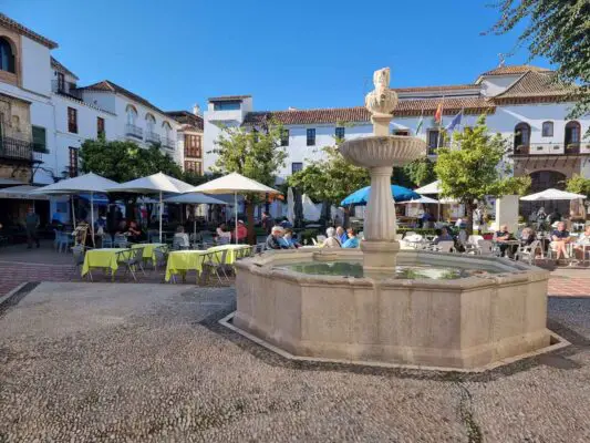 A round stone fountain in Plaza de los Naranjos in the centre of the Marbellaold town. The square is small and it is lined with orange trees in front of the white buildings. In the middle there are cafes and restaurants with outdoor terraces.